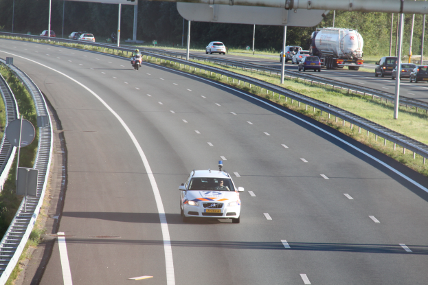 Ein Auto fährt auf einer Autobahn mit einem Polizeiwagen am Straßenrand, anderen Fahrzeugen, Geländern, Pfählen, Planken und Bäumen im Hintergrund.