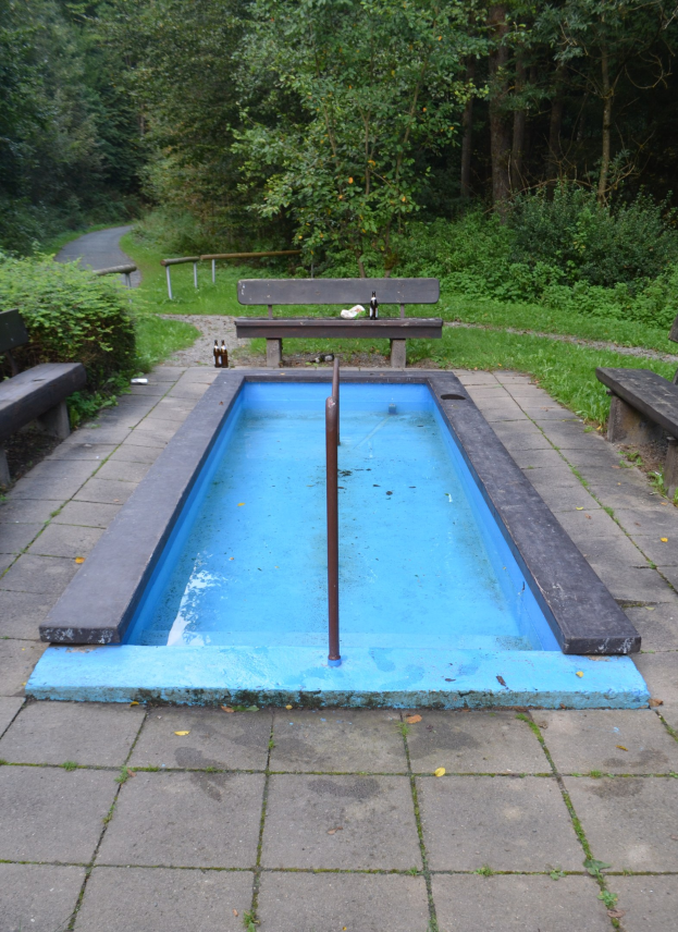 Kleiner Parkpool mit zentralem Pfahl, umgeben von Bänken, Grünfläche, Straße und Bäumen.