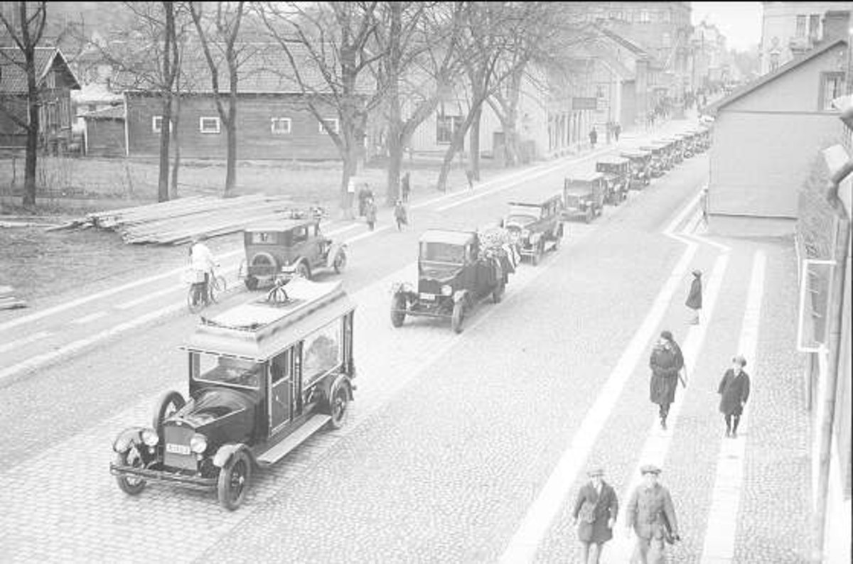 Schwarze und weiße Straßenszene in Berlin aus den 1930er-1940er Jahren, die alte Autos, Fußgänger, Gebäude mit Fenstern, Bäume und einen klaren Himmel zeigt.
