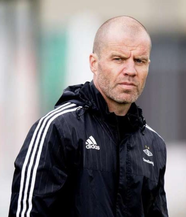 Bald coach in einer marineblauen Jacke mit weißen Streifen und weißem Kragen, der auf einem Fußballfeld steht und die Hände gefaltet hat.