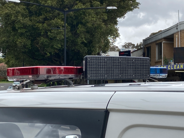 Ein Polizeiwagen mit eingeschaltetem Blaulicht steht an der Straße neben Gebäuden, Bäumen, Pfählen und Schildern mit Text, unter einem bewölkten Himmel, wahrscheinlich nach einem tödlichen Schusswechsel an einer Schule in Brisbane.