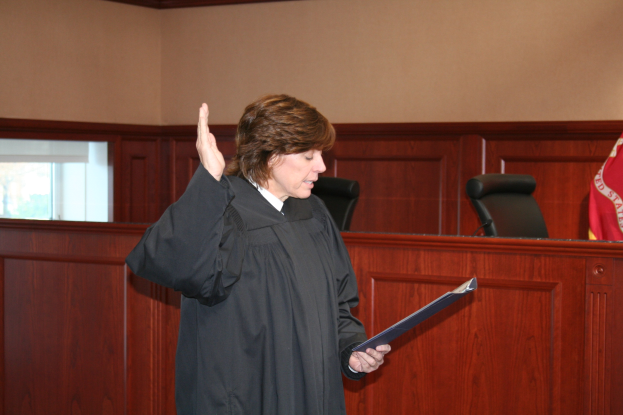 Eine Frau in einer schwarzen Robe steht an einem Richtertisch und hält eine Akte, mit einem Mikrofon und einer Flagge auf dem Tisch, Stühle und ein Fenster dahinter.