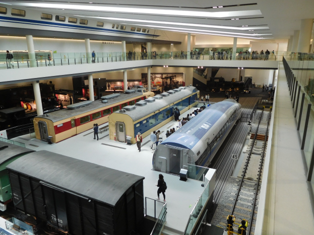 Eine geräumige Museumsausstellung zeigt verschiedene Züge in unterschiedlichen Größen und Farben, bei der Besucher von verschiedenen Ebenen und Stockwerken aus zuschauen.