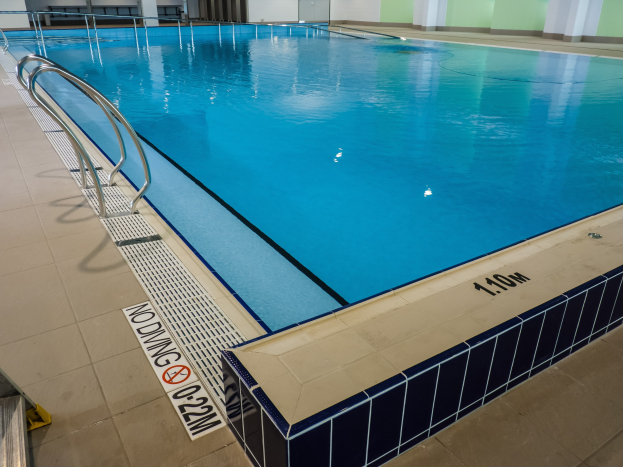 Großes Hallenbad in einem geräumigen Gebäude mit gefliestem Boden, Metallträgern, einer Tafel, einer Tür, einer Wand und Deckenleuchten.