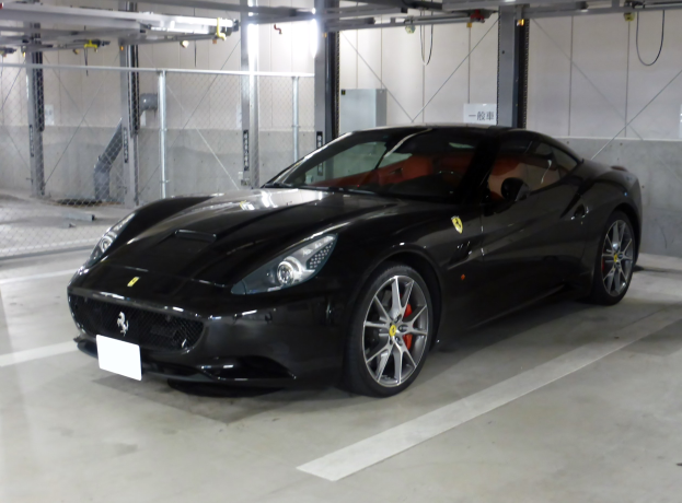 Schwarzer Ferrari California in einer Parkgarage mit einer Wand im Hintergrund, Maschendraht auf der linken Seite und Deckenbeleuchtung.