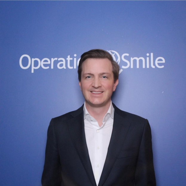 Ein Mann in einem Anzug, der vor einer blauen Wand mit der Aufschrift "Operationsmile" lächelt