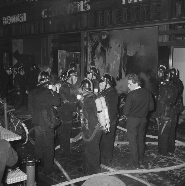 Schwarzes und weißes Bild von Feuerwehrleuten in Helmen mit Sauerstoffflaschen, die vor einem Gebäude stehen, mit einem Fahrrad und einem Tisch mit Gegenständen in der Nähe.