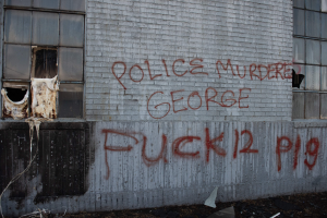 Ein Gebäude mit Graffiti an der Seite, das "Polizei ermordet George Fuck 12 P19" lautet, mit Fenstern und diversen Gegenständen vor dem Gebäude.