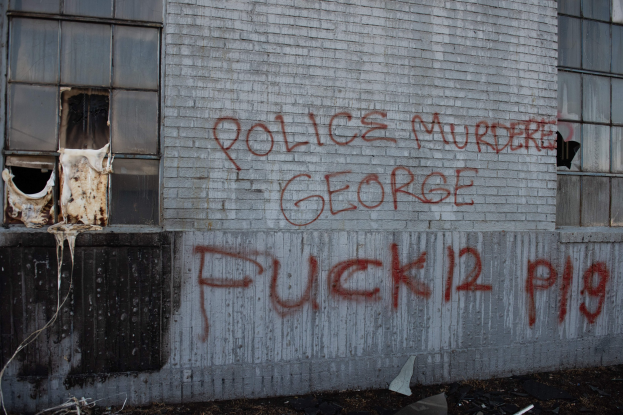 Ein Gebäude mit Graffiti an der Seite, das "Polizei ermordet George Fuck 12 P19" lautet, mit Fenstern und diversen Gegenständen vor dem Gebäude.