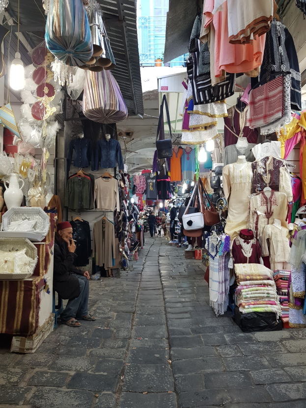Ein belebter Straßenmarkt in der Altstadt von Jerusalem mit Ständen, die Kleidung, Taschen, Lampen und andere Gegenstände verkaufen, während Menschen umhergehen und die Stände durchstöbern, mit einem Gebäude im Hintergrund.