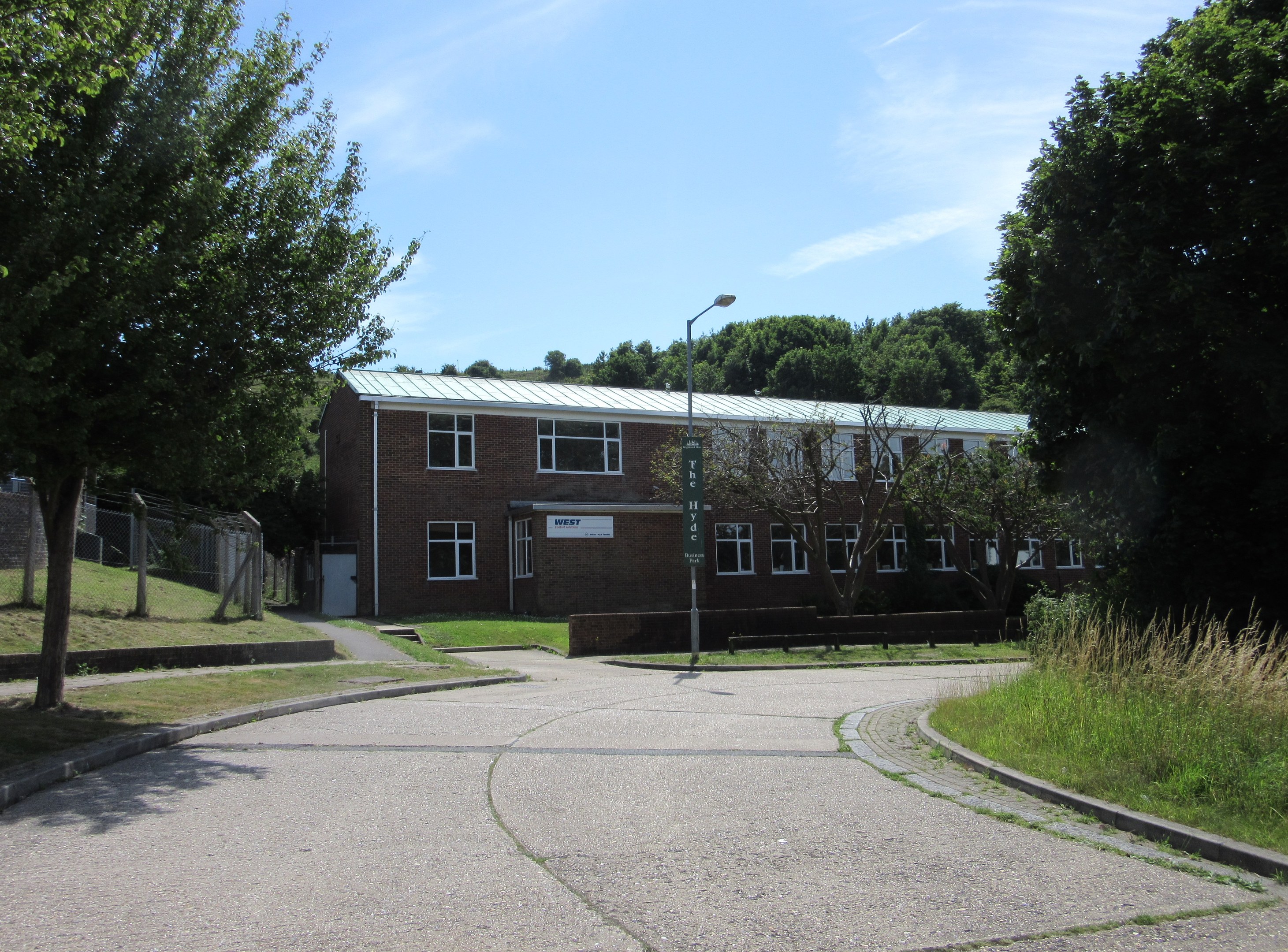 Ein Schulgebäude mit Fenstern, einem Straßenpfahl, einem Schild, einer Baumgruppe, Gras, einem Zaun und einem bewölkten Himmel, das sich in einem Park mit saftigem Gras befindet.