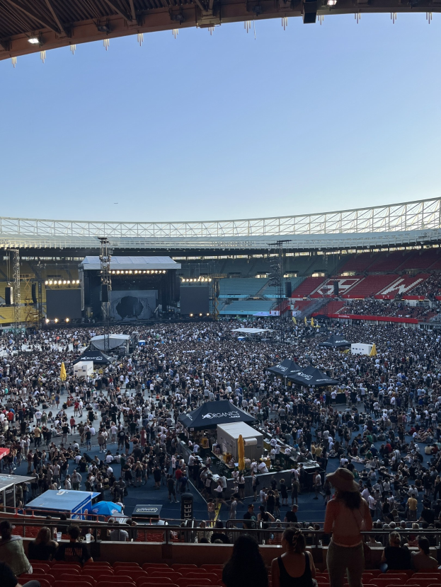 Große Menge von Menschen auf Sitzplätzen und Stehplätzen in einem Stadion mit Zelten, Geländern und Deckenlampen unter einer Dachkonstruktion, mit dem Himmel im Hintergrund.