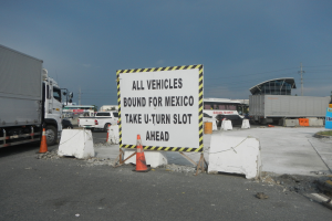 Ein Lkw auf einem Parkplatz neben einem Schild mit der Aufschrift "Alle Fahrzeuge nach Mexiko". Im Hintergrund sind Gebäude, Bäume, Pfosten und ein klarer blauer Himmel zu sehen.