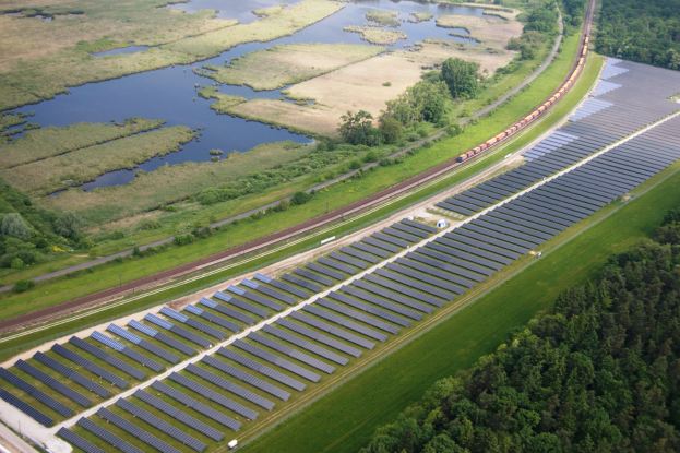 Luftaufnahme einer Solaranlage mit Solarmodulen auf einem Feld, umgeben von Bäumen, Gras, Pflanzen und Wasser, mit einem Zug auf einer nahen Bahnstrecke.