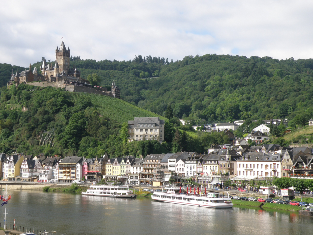Ein malerischer Blick auf den Rhein in Deutschland mit einer Burg auf einem Hügel, Booten auf dem Fluss, Fahrzeugen auf der Straße und einem bewölkten Himmel.