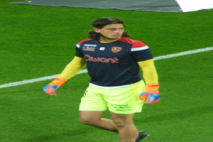 Ein Mann in einem blauen Hemd und gelben Shorts läuft auf einem Fußballfeld, die Arme ausgestreckt und mit entschlossener Miene, mit einem Tuch oben auf dem Bild.