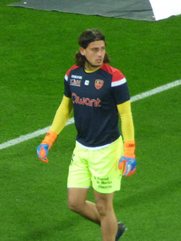 Ein Mann in einem blauen Hemd und gelben Shorts läuft auf einem Fußballfeld, die Arme ausgestreckt und mit entschlossener Miene, mit einem Tuch oben auf dem Bild.