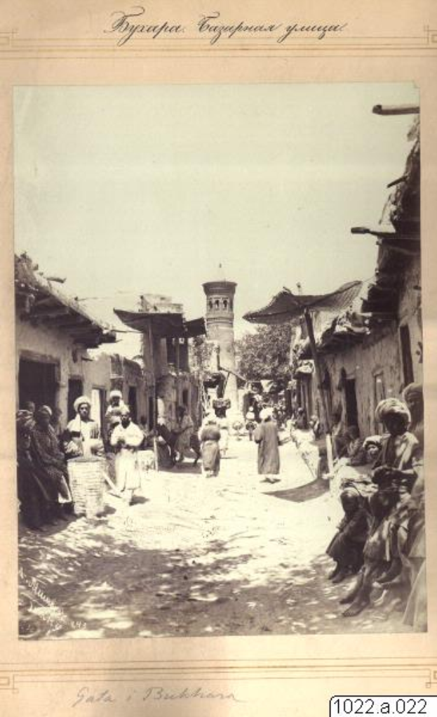 Ein altes Schwarz-Weiß-Foto von Menschen, die eine Straße in einem kleinen Ort entlanggehen, mit Häusern auf beiden Seiten und einem Turm im Hintergrund, mit Text oben und unten auf dem Bild.