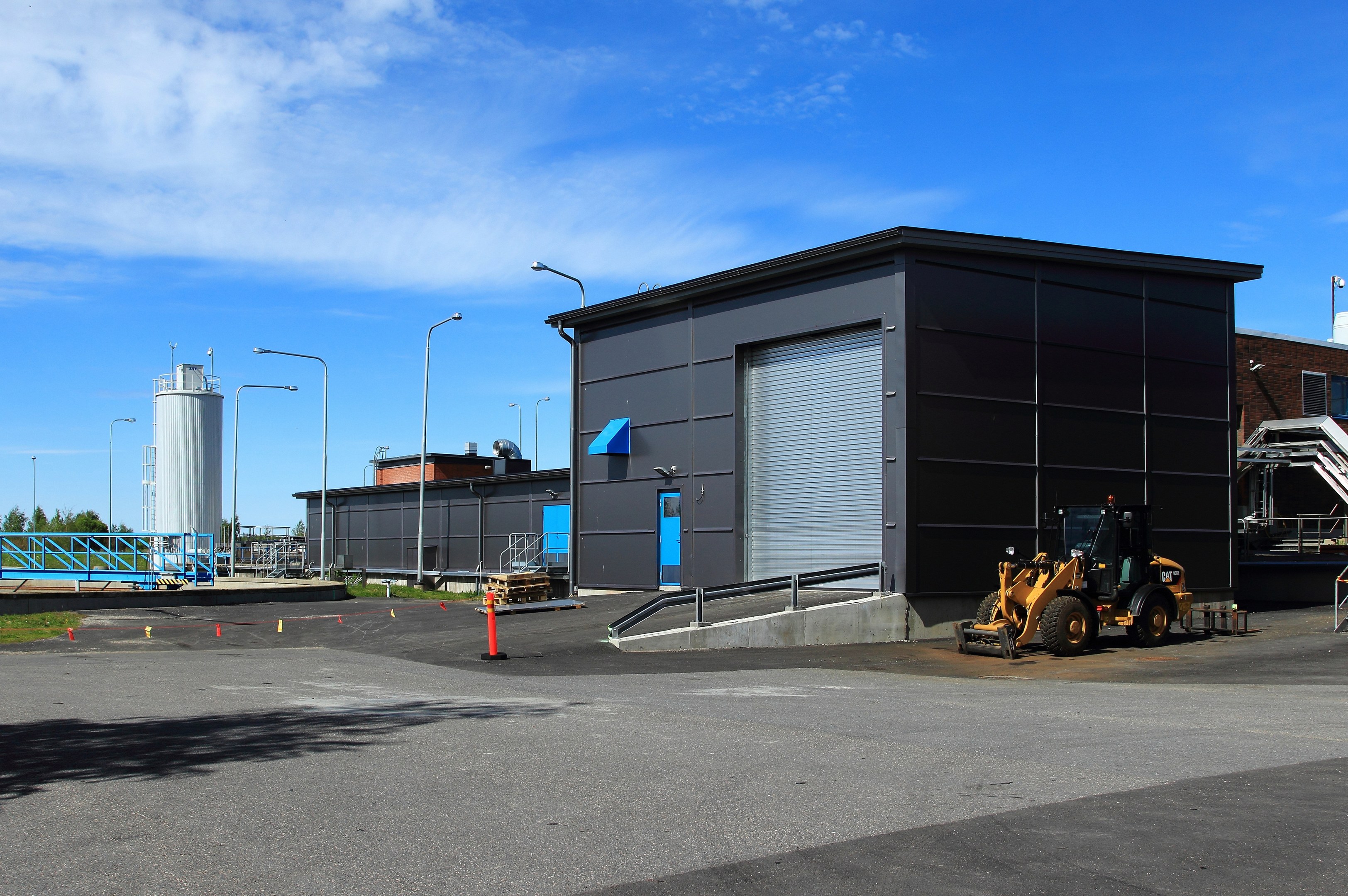 Große Industriehalle mit einem Bagger davor, umgeben von Straßeninfrastruktur und Fahrzeugen, unter einem bewölktem Himmel.