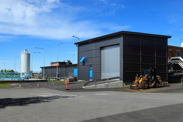 Große Industriehalle mit einem Bagger davor, umgeben von Straßeninfrastruktur und Fahrzeugen, unter einem bewölktem Himmel.