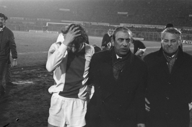 Eine Gruppe Männer in Not steht zusammen auf einem Fußballfeld, mit einem Stadion im Hintergrund, auf einem Schwarz-Weiß-Bild.
