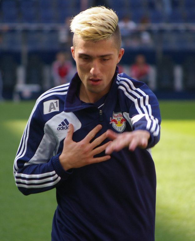 Ein junger Mann in einem blauen T-Shirt steht auf einem Fußballfeld und streckt die Arme aus, mit einem entschlossenen Gesichtsausdruck und einem unscharfen Hintergrund.