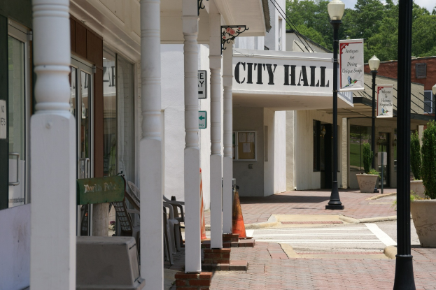 Rathaus mit umliegenden Gebäuden, Straßeninfrastruktur, Kraftfahrzeugen, Verkehrsleitelementen und Grünflächen unter einem bewölkten Himmel.
