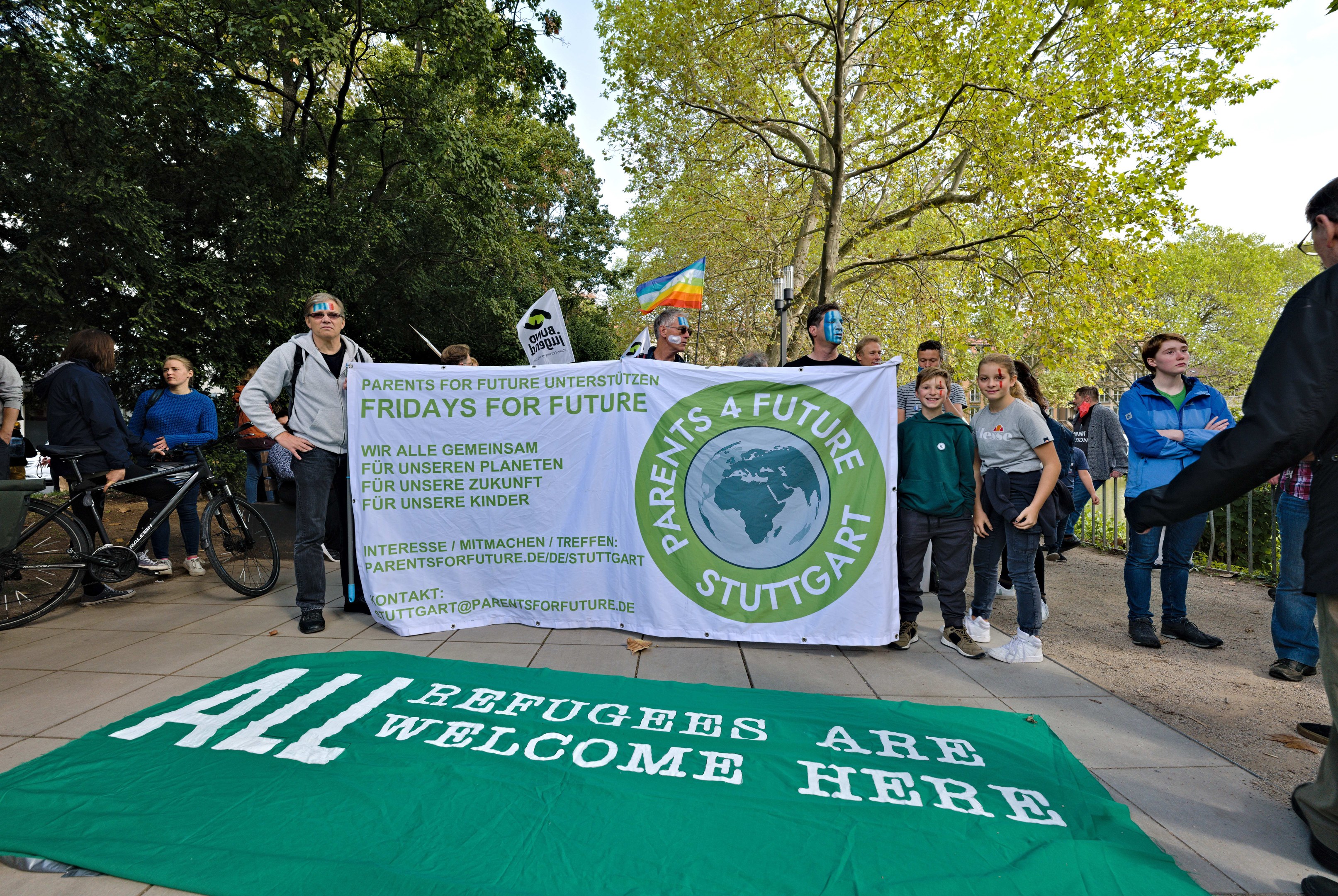 Eine Gruppe von Menschen mit einem Banner mit der Aufschrift "All Refugees Are Welcome Here" und einer Flagge, mit Fahrrädern, einem Zaun, einem Straßenschild, einer Tafel, Bäumen und einem bewölkten Himmel im Hintergrund.
