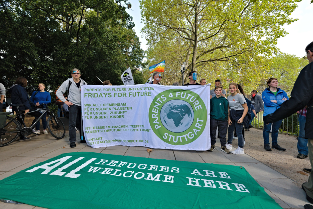 Eine Gruppe von Menschen mit einem Banner mit der Aufschrift "All Refugees Are Welcome Here" und einer Flagge, mit Fahrrädern, einem Zaun, einem Straßenschild, einer Tafel, Bäumen und einem bewölkten Himmel im Hintergrund.