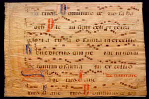 Ein illuminiertes Manuskript mit Notenschrift auf einem Blatt Papier vor dunklem Hintergrund, gefüllt mit Text und Musiksymbolen, die ein komplexes Design bilden.