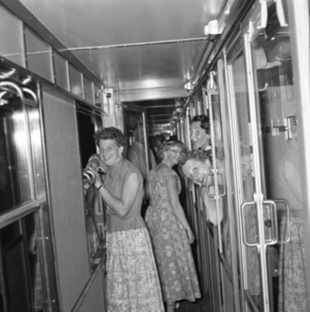 Eine Gruppe von Frauen auf einem Zug mit Türen auf beiden Seiten und Lampen oben, dargestellt in Schwarz-Weiß.