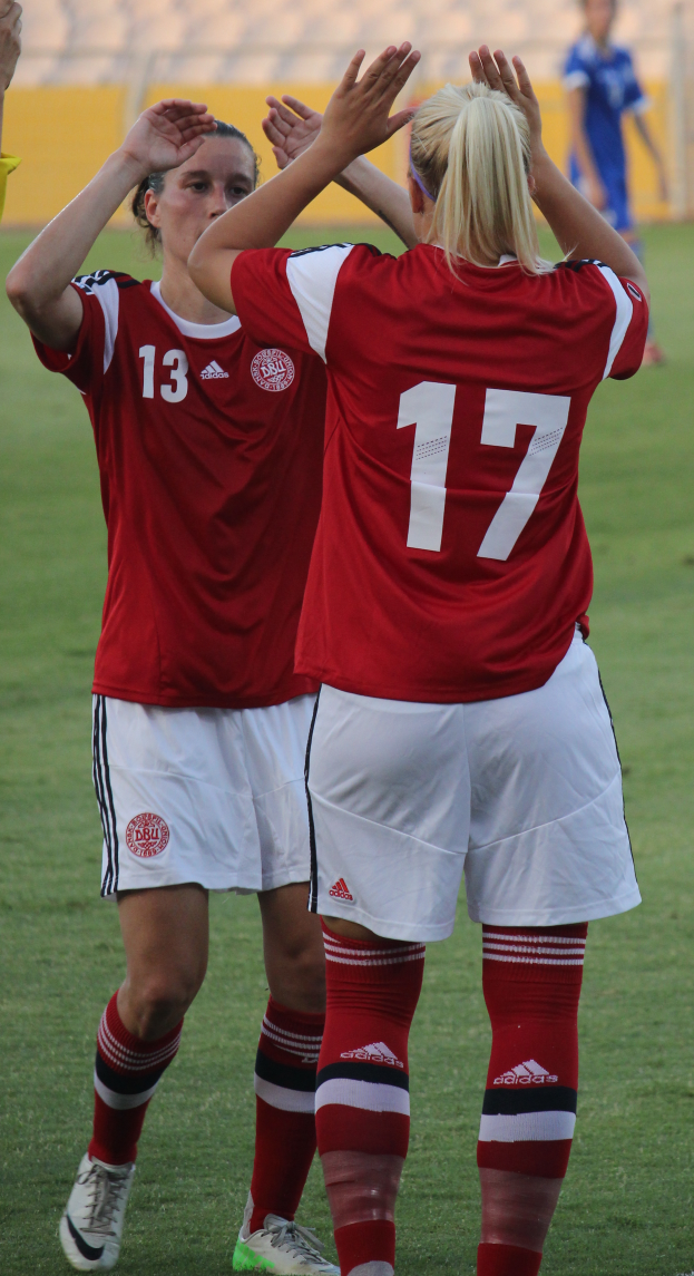 Zwei Frauen in roten T-Shirts und weißen Shorts stehen auf einem Fußballfeld und klatschen in die Hände, im Hintergrund ist eine Person in einem blauen Kleid, eine gelbe Tafel und ein paar Stühle zu sehen.
