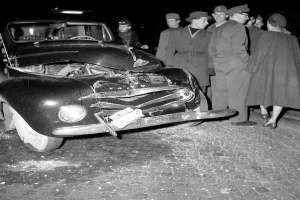 Ein Schwarz-Weiß-Bild eines auf der Straße liegenden Wagens mit eingedellter Motorhaube und zersplitterten Fenstern, umgeben von einer Gruppe besorgter Menschen.