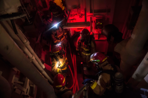 Eine Gruppe von Feuerwehrmännern in Helmen und Sauerstoffzylindern steht in einem dunklen Raum mit Rohren auf beiden Seiten, mit einer Maschine und anderen Gegenständen im Hintergrund.