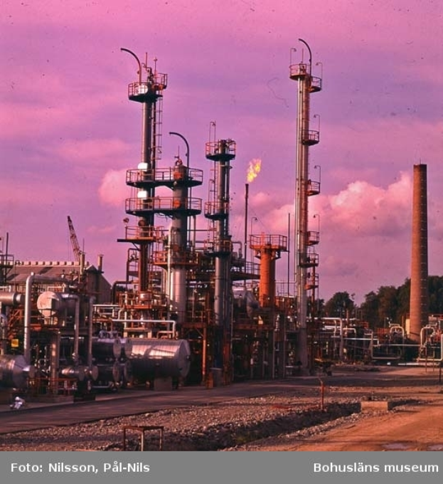 Eine Ölfabrik mit zahlreichen Rohren, Ventilen, Metallkonstruktionen, Containern, einem Gebäude, einem Kran und einer Gruppe von Bäumen unter einem bewölkten Himmel, mit Text am unteren Bildrand.