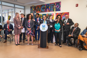Eine Gruppe von Menschen, die in einem Raum mit einem Podium, einem Mikrofon und einer Tafel mit der Aufschrift "Sancho City Councilman Joins City Council" stehen und sitzen, mit Flaggen, Feuerlöscher und Deckenleuchten an der Rückwand.