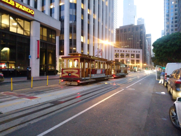 Eine Seilbahn fährt eine Straße in San Francisco entlang mit Fahrzeugen, einem Fußgänger und einem geparkten Van, eingerahmt von Gebäuden, Bäumen und Himmel.