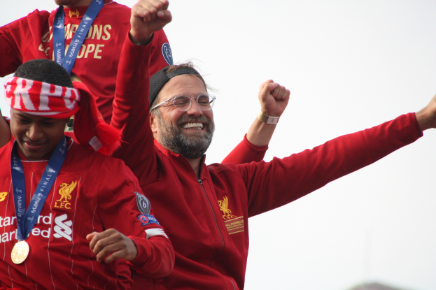 Liverpool-Trainer Jürgen Klopp, der eine rote Jacke trägt, feiert mit seinen Spielern nach dem Gewinn der UEFA Champions League, während zwei medaillentragende Personen in der Nähe sind und der Himmel im Hintergrund zu sehen ist.