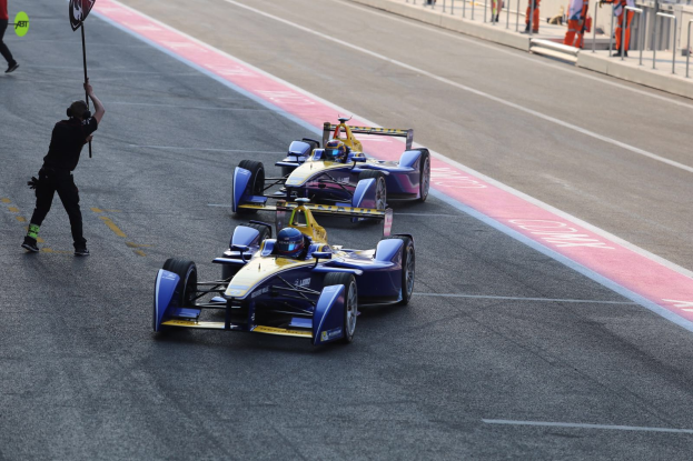 Zwei Formel-E-Autos auf einer Rennstrecke mit einem Fahne schwingenden Mann in der Nähe, während einige Menschen links gehen und andere im Hintergrund hinter Gittern sichtbar sind.