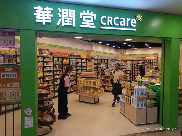 Eine Gruppe von Menschen vor einem Geschäft mit einem "crcare"-Schild, in dem sich Regale mit Lebensmitteln, Flaschen und anderen Gegenständen befinden, mit Lampen an der Decke und einer Tafel auf der linken Seite, die darauf hinweist, dass es Teil einer chinesischen Supermarktkette ist, die ihren ersten Online-Shop eröffnet.