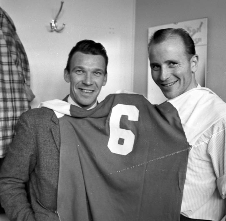 Zwei Männer in Schwarz-Weiß, die strahlend eine Fußballtrikot hochhalten, mit einer Tafel und einem Kleiderbügel an der Wand dahinter.