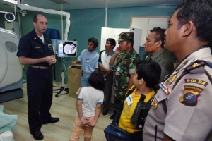 Eine Gruppe von Menschen steht in einem Krankenhauszimmer, ein Mann hält ein Mikrofon in der Mitte, ein Bett mit einer Decke darauf links und verschiedene medizinische Geräte, ein Röntgengerät, eine Tafel mit Text, eine Tür, eine Wand und eine Decke mit Deckenleuchten im Hintergrund.