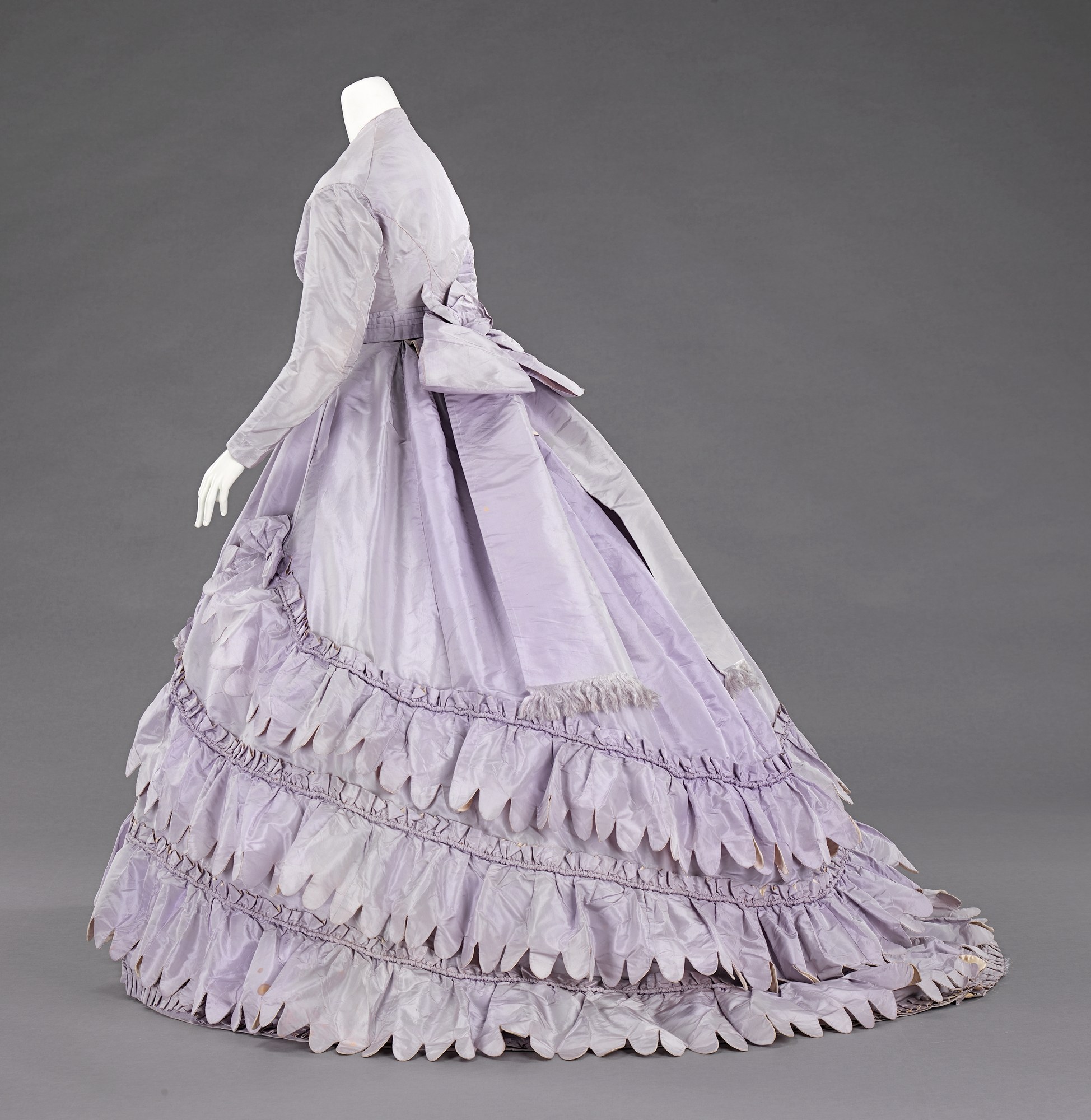 Eine Schaufensterpuppe in einem lila Ballkleid mit engem Oberteil, vollem gerüschtem Rock und langen Ärmeln vor einem grauen Hintergrund.