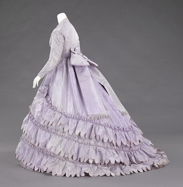 Eine Schaufensterpuppe in einem lila Ballkleid mit engem Oberteil, vollem gerüschtem Rock und langen Ärmeln vor einem grauen Hintergrund.