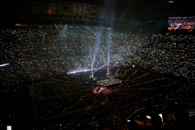 Eine große Menschenmenge in einem Stadion bei Nacht, beleuchtet von hellen Lichtern, mit einem Bildschirm auf der rechten Seite, der wahrscheinlich die Ergebnisse der Super Bowl XLVII-Halbzeitpause anzeigt.