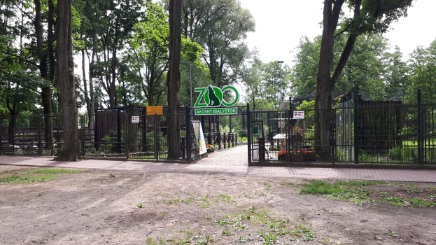Eintritt zu einem Zoo mit einem "Zoo"-Schild, umgeben von einem Metallzaun, Bäumen, Blumenpflanzen, einem Straßenpfahl und einem bewölkten Himmel.
