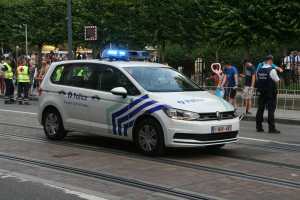 Ein Polizeiwagen fährt auf einer Straße neben einer Menschenmenge, einige tragen Mützen, mit einem Geländer, Bäumen und Laternen im Hintergrund.