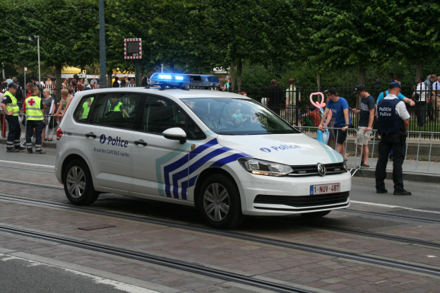 Ein Polizeiwagen fährt auf einer Straße neben einer Menschenmenge, einige tragen Mützen, mit einem Geländer, Bäumen und Laternen im Hintergrund.