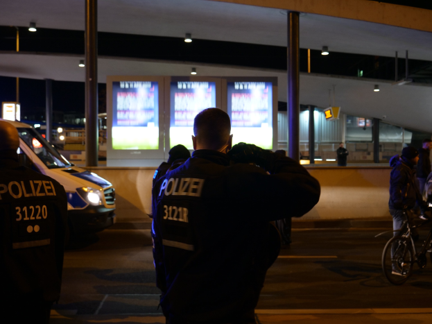 Eine Gruppe von Polizeibeamten steht vor einem hell erleuchteten Gebäude in der Nacht, mit einem geparkten Auto auf der linken Seite und einem Fahrradfahrer auf der rechten Seite.