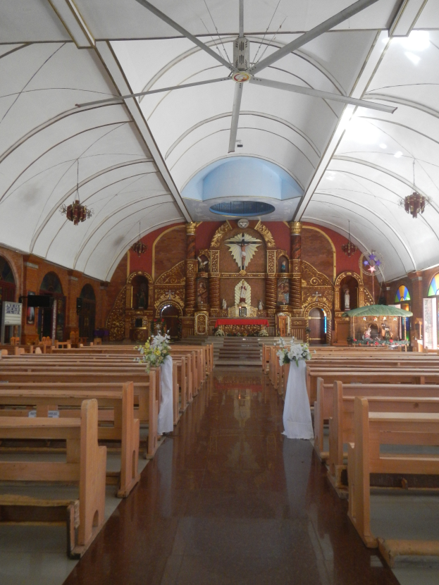 Das Innere einer Kirche mit hölzernen Kirchenbänken, Blumensträußen auf dem Boden, Deckenlampen, Buntglasfenstern auf beiden Seiten und Skulpturen an den Wänden im Hintergrund.